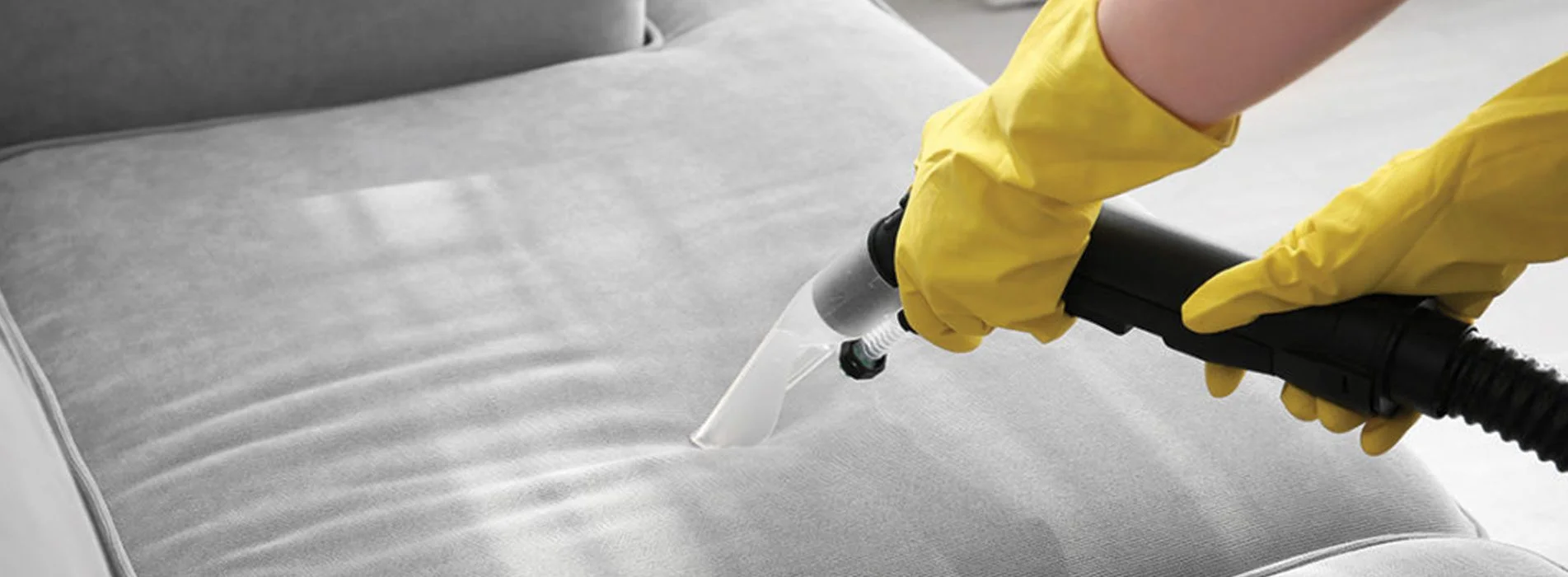 A person in yellow jacket using a spray gun to clean white couch.